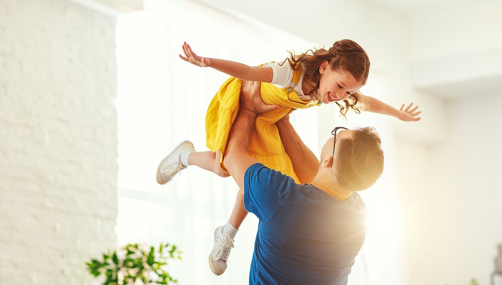 Das Bild zeigt einen Erwachsenen, der ein lächelndes Kind in einem gelben Kleid hochhebt. Das Kind streckt die Arme seitlich aus, als würde es fliegen. Die Szene findet in einem hellen, sonnendurchfluteten Raum statt und vermittelt Freude und Nähe.