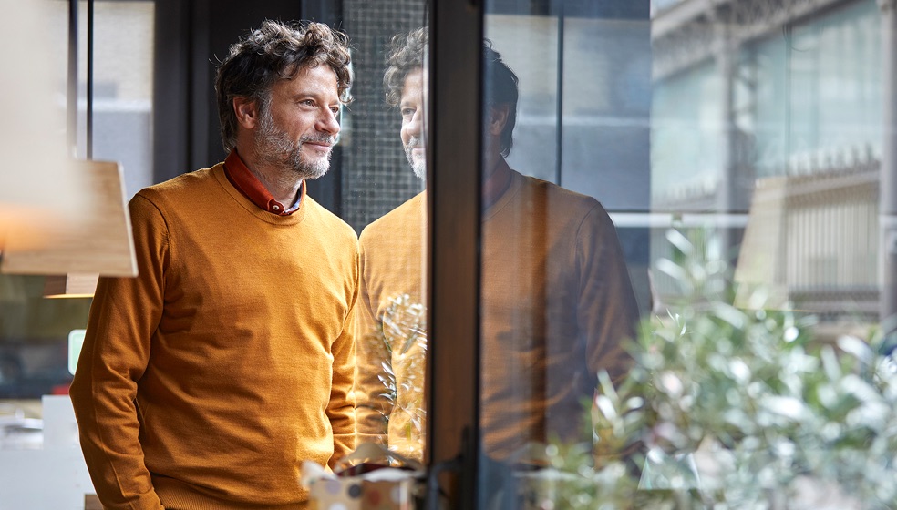 Mann mit lockigen braunen kurzen Haaren, Brille und gelbem Pullover steht vor einem Glasfenster und blickt nachdenklich nach draußen