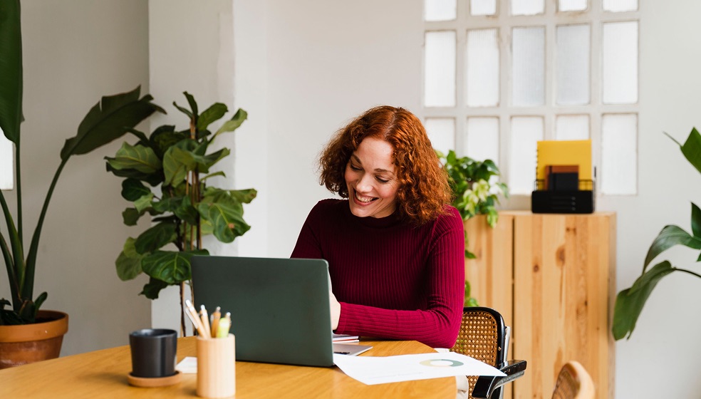 Frau mit halblangen roten Locken sitzt mit ihrem Notebook an einem Schreibtisch und blickt lächelnd auf den Bildschirm