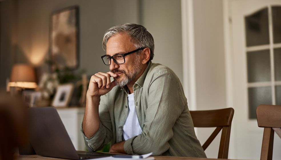Eine Person mit grauen Haaren sitzt an einem Tisch vor einem aufgeklappten Laptop in einem wohnlich eingerichteten Raum. Im Hintergrund sind ein Fenster, ein Regal mit Bildern und eine Lampe zu sehen. Die Person trägt eine Brille sowie ein helles Hemd über einem T-Shirt.