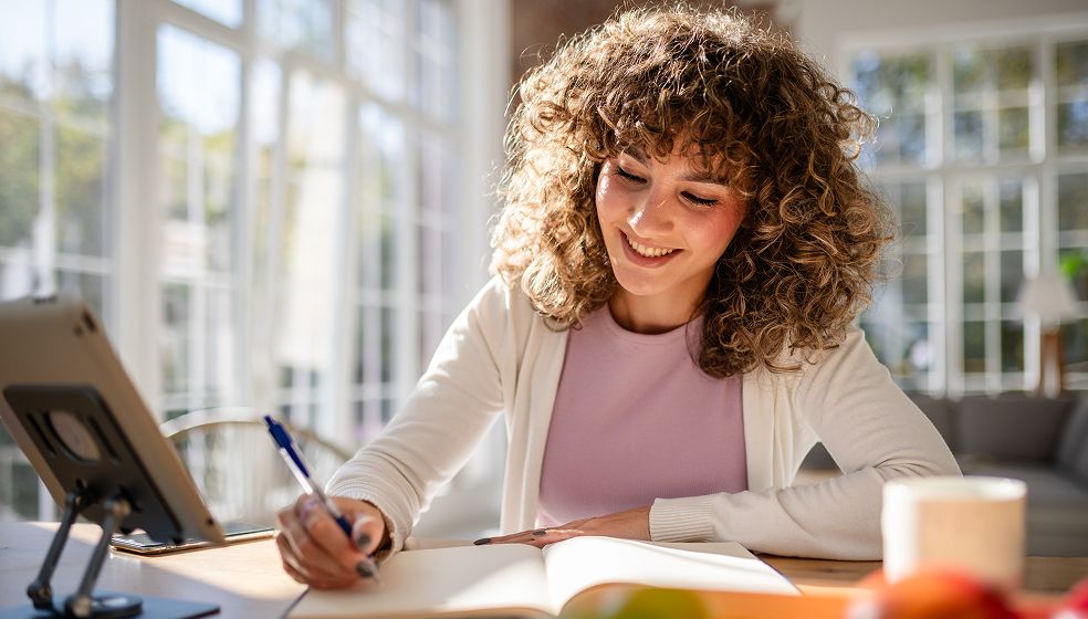 Eine Person mit lockigem Haar sitzt an einem Tisch in einem hellen Raum. Sie schreibt in ein geöffnetes Heft oder Notizbuch und hat einen Stift in der Hand. Vor ihr steht ein Tablet auf einem Ständer. Im Vordergrund befindet sich eine Tasse. Im Hintergrund sind große Fenster mit viel Tageslicht zu sehen.