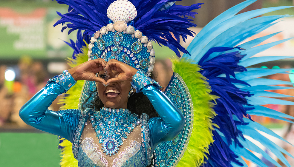 Das Bild zeigt eine Person in einem farbenfrohen Kostüm mit einem aufwendigen Kopfschmuck und Flügeln. Das Kostüm ist überwiegend in Blau- und Türkistönen gehalten, mit vielen Glitzersteinen, Perlen und Verzierungen. Große blaue und grüne Federn schmücken den Rücken und Kopf der Person. Die Person trägt langärmelige Kleidung und hebt beide Hände an den Kopf. Der Hintergrund ist unscharf und grünlich.