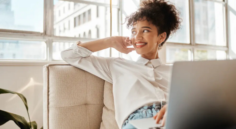 Frau im dunklen Afro in weißer Bluse hat ein Notebook auf dem Schoß und blickt lächelnd zur Seite