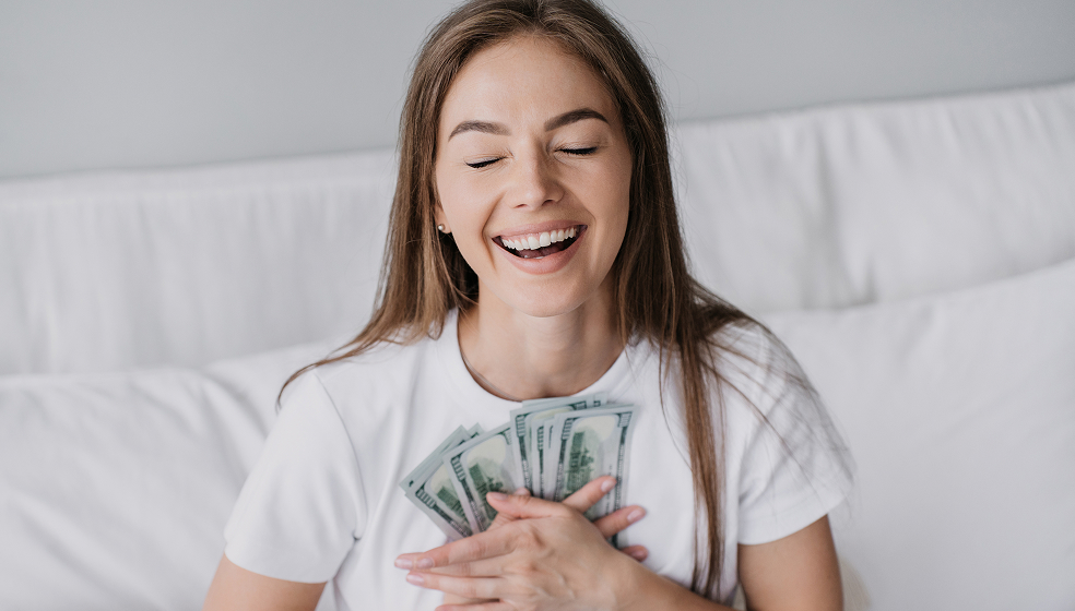 Eine Person mit langen braunen Haaren sitzt auf einem weißen Sofa oder Bett. Sie trägt ein weißes T-Shirt und hält mehrere US-Dollar-Geldscheine mit beiden Händen vor der Brust.