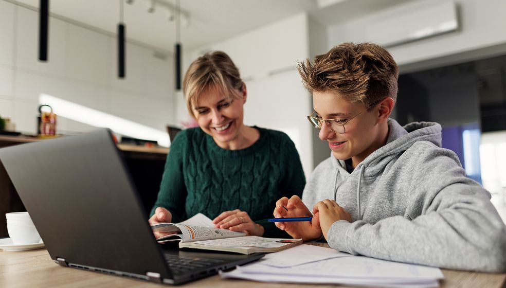 Die Abbildung zeigt eine erwachsene Person und ein Kind, die gemeinsam an einem Tisch sitzen. Vor ihnen liegen ein geöffnetes Buch, Notizblätter und ein Laptop. Die Umgebung wirkt wie ein modernes Zuhause. Es sieht nach gemeinsamer Lern- oder Hausaufgabenzeit aus.