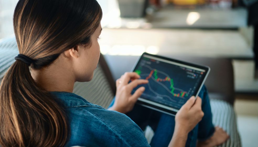 Eine Person sitzt auf einem Sofa und hält ein Tablet in den Händen. Auf dem Bildschirm ist ein Diagramm mit Kurven und Balken zu sehen, vermutlich ein Finanz- oder Börsendiagramm mit rot-grünen Kursverläufen.