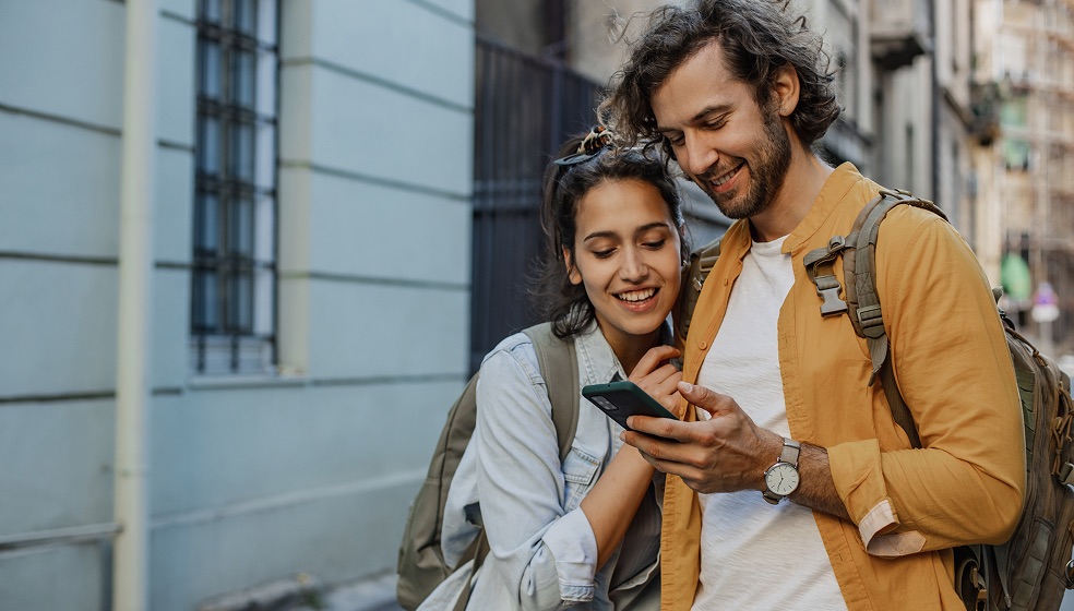 Das Bild zeigt zwei Personen mit Rucksäcken, die in einer städtischen Umgebung nebeneinander stehen. Eine Person hält ein Smartphone, auf das beide gemeinsam schauen. Beide tragen Freizeitkleidung. Im Hintergrund sind Häuserfassaden und ein Gehweg zu sehen.