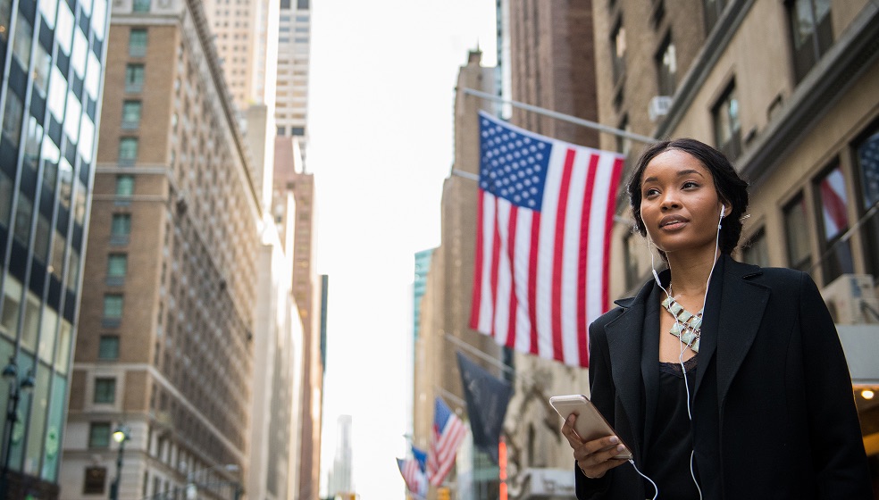 Eine Person steht in einer Großstadtstraße zwischen hohen Gebäuden, hält ein Smartphone in der Hand und trägt Kopfhörer. Im Hintergrund sind mehrere große US-amerikanische Flaggen an den Gebäuden befestigt.