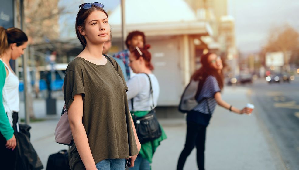 Das Bild zeigt mehrere Personen, die an einer Bushaltestelle im städtischen Raum auf den Bus warten. Im Vordergrund steht eine Frau mit Sonnenbrille auf dem Kopf und einer Umhängetasche. Im Hintergrund sind weitere wartende Personen zu erkennen. Die Umgebung wirkt urban, mit Straßen und Gebäuden im Hintergrund.