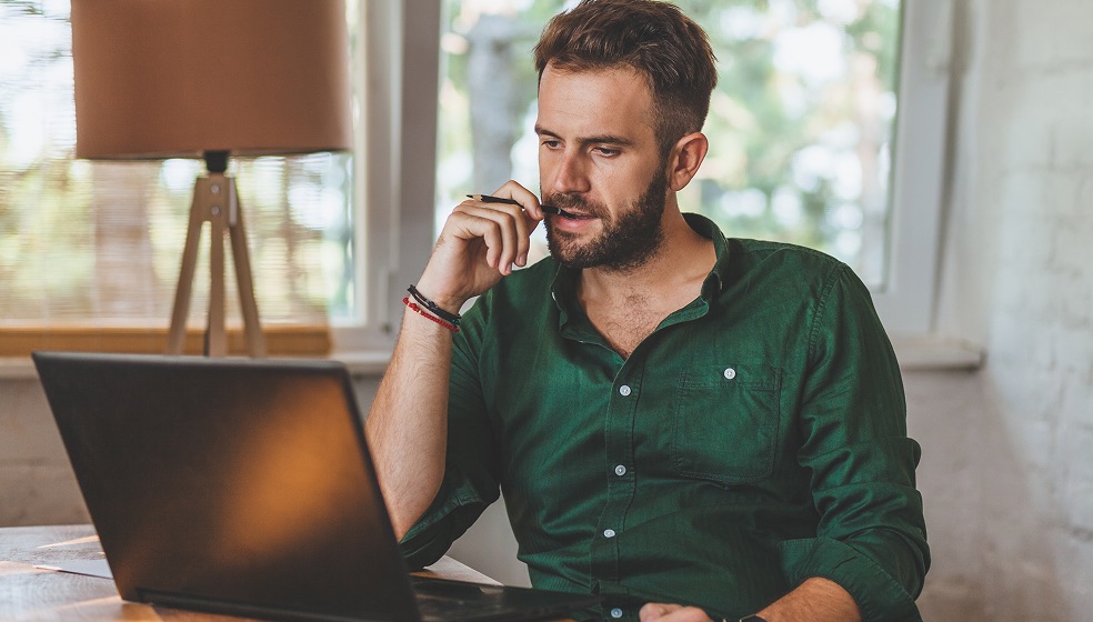 Mann mit kurzen blonden Haaren und grünem Hemd sitzt an einem Schreibtisch und blickt nachdenklich auf seinen Laptop. 