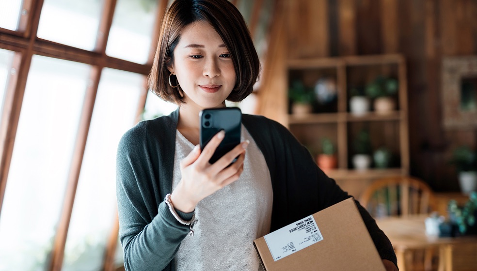 Frau mit halblangen dunkelbraunen Haaren blickt lächenld auf ihr Smartphone. Unter dem Arm hält sie ein Paket.