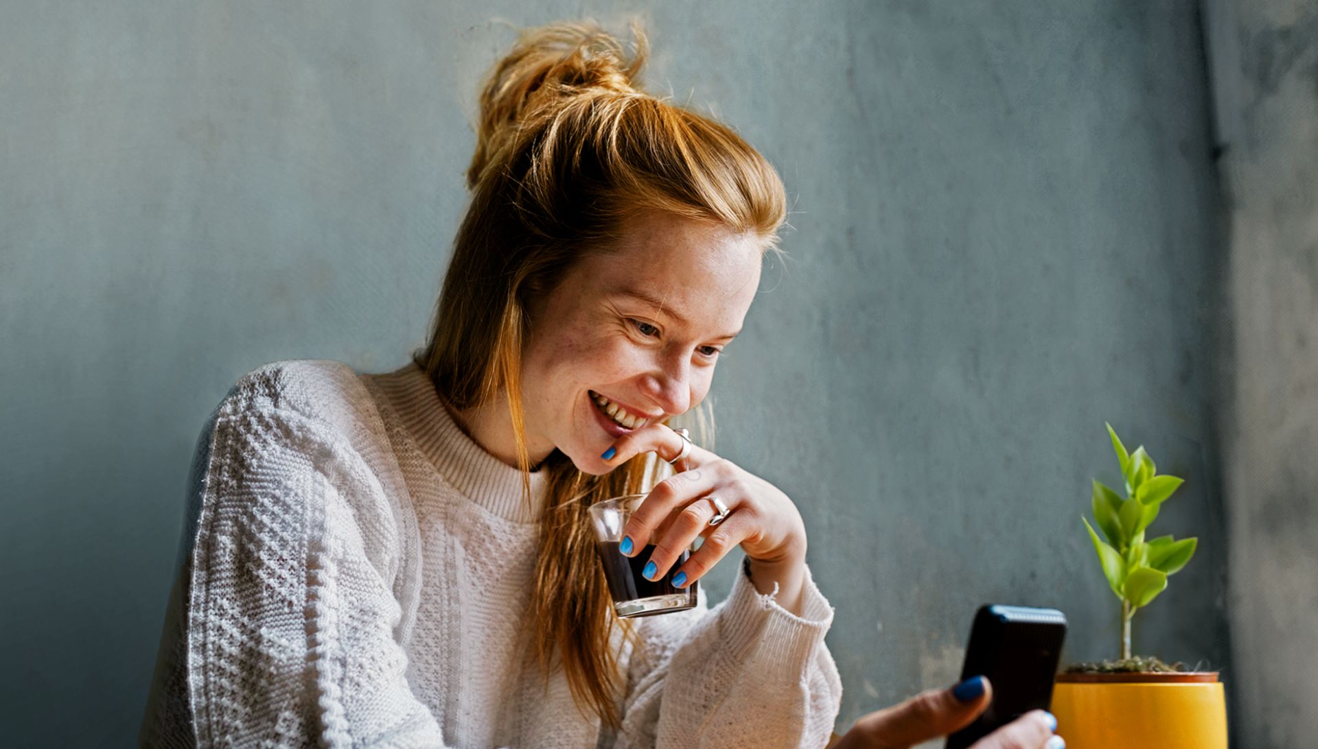 Das Bild zeigt eine Person mit langen, rötlichen Haaren, die einen hellen Strickpullover trägt. Sie sitzt an einem Tisch, hält in der einen Hand ein Glas mit einem dunklen Getränk und in der anderen Hand ein Smartphone. Auf dem Tisch steht rechts eine kleine grüne Pflanze in einem gelben Blumentopf. Der Hintergrund ist eine schlichte graue Wand.