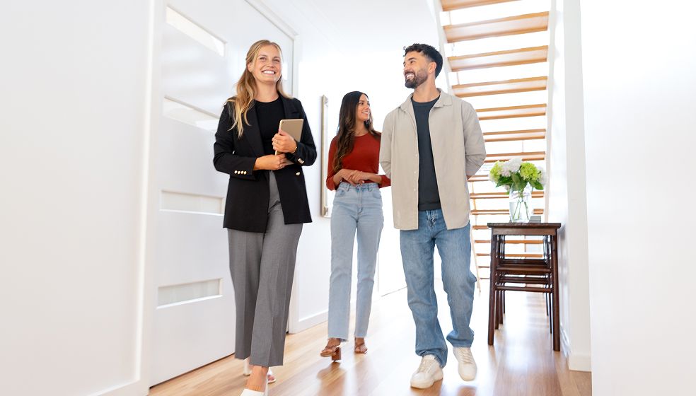 Das Bild zeigt drei Personen, die gemeinsam durch einen hellen Flur mit Holzfußboden gehen. Eine Person hält ein Tablet in der Hand, die anderen beiden hören zu. Im Hintergrund steht ein kleiner Tisch mit einem Blumenstrauß. Die Szene wirkt freundlich und modern, möglicherweise handelt es sich um eine Immobilienbesichtigung.