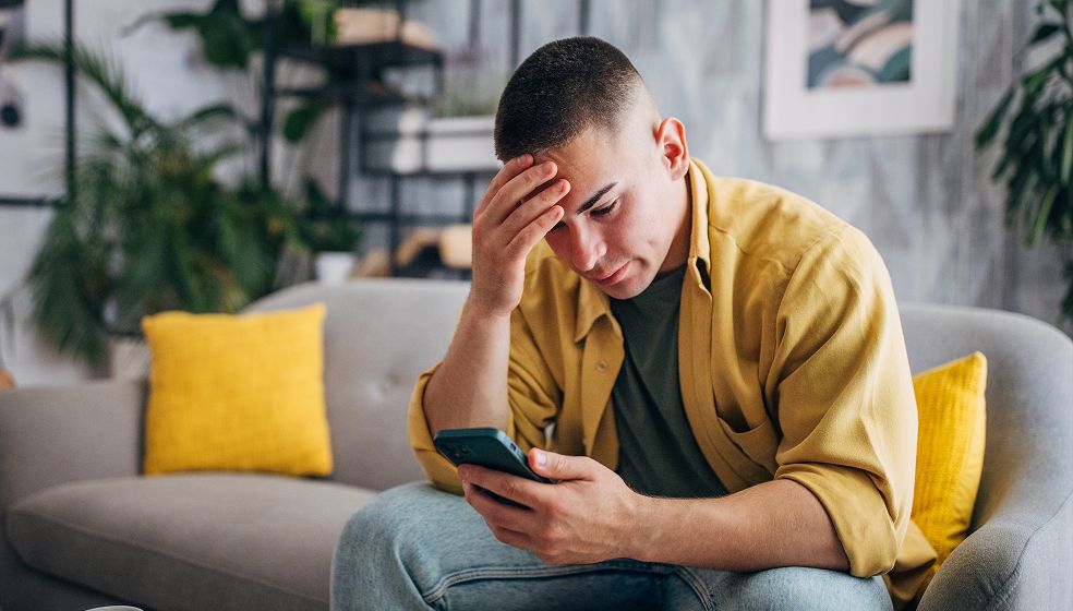 Ein Mann sitzt auf einem Sofa in einem Wohnzimmer. Er hält ein Smartphone in der linken Hand und stützt sich mit der rechten Hand an den Kopf. Im Hintergrund sind Pflanzen, Regale und ein Bild an der Wand zu sehen. Auf dem Sofa liegen gelbe Kissen. Die Stimmung wirkt nachdenklich oder angespannt.