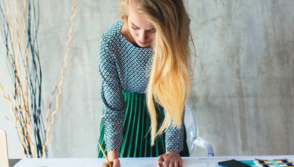 Blonde Frau mit Kleid steht an einem Schreibtisch voller Unterlagen und schreibt etwas auf