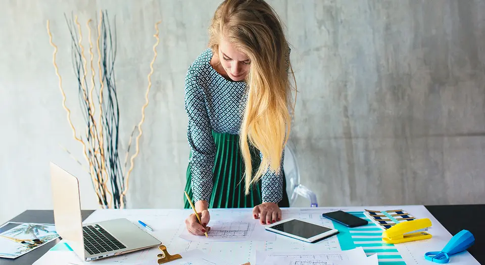 Blonde Frau mit Kleid steht an einem Schreibtisch voller Unterlagen und schreibt etwas auf