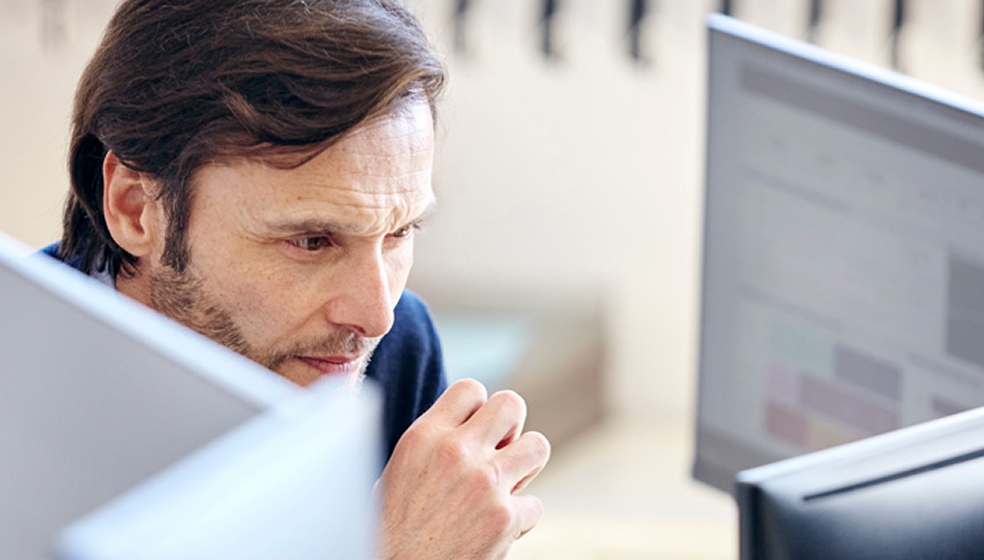 Dunkelhaariger Mann sitzt vor mehreren Bildschirmen und schaut konzentriert hinein