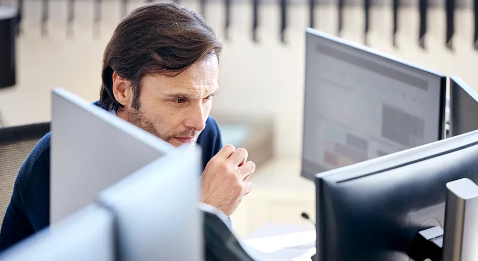 Dunkelhaariger Mann sitzt vor mehreren Bildschirmen und schaut konzentriert hinein