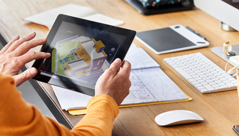 Mann in orangenem Pulli sitzt an einem Schreibtisch schaut in sein Tablet