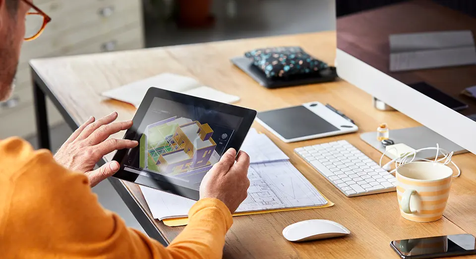 Mann in orangenem Pulli sitzt an einem Schreibtisch schaut in sein Tablet