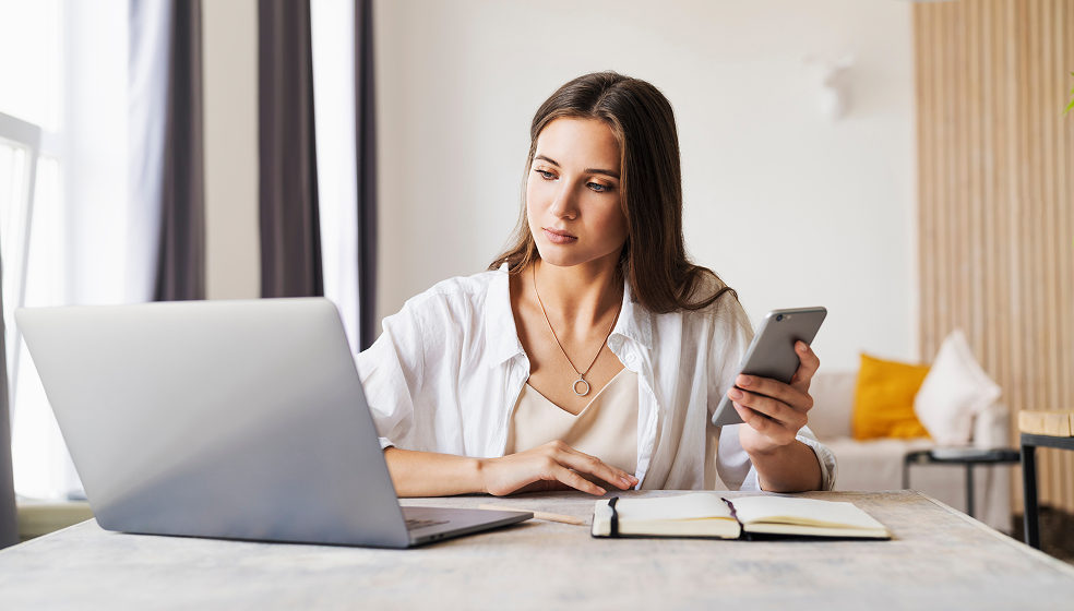 Frau in weißem Hemd arbeitet am Schreibtisch und hält ihr Smartphone in der Hand