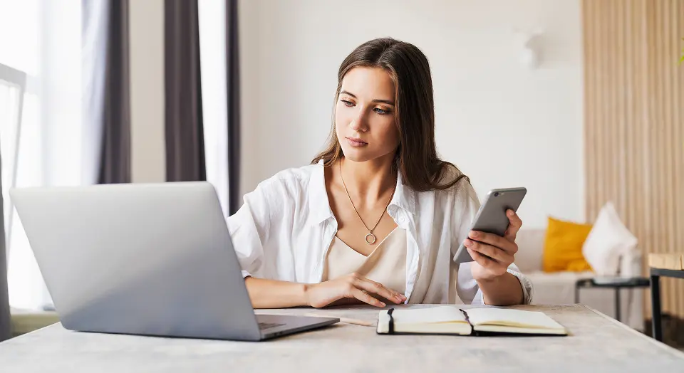 Frau in weißem Hemd arbeitet am Schreibtisch und hält ihr Smartphone in der Hand