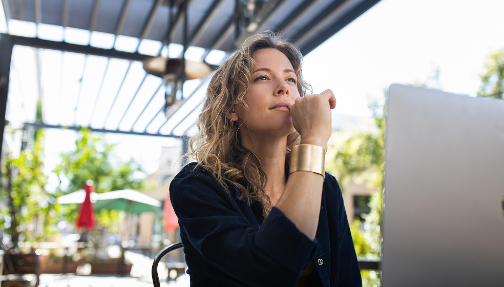 Blonde Frau in schwarzem Hemd arbeitet im Garten am Laptop und schaut grübelnd in die Ferne