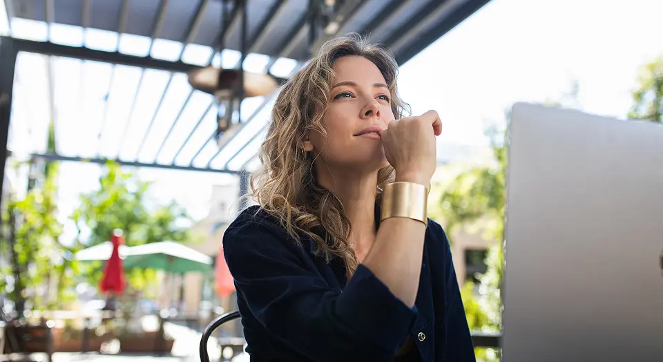 Blonde Frau in schwarzem Hemd arbeitet im Garten am Laptop und schaut grübelnd in die Ferne