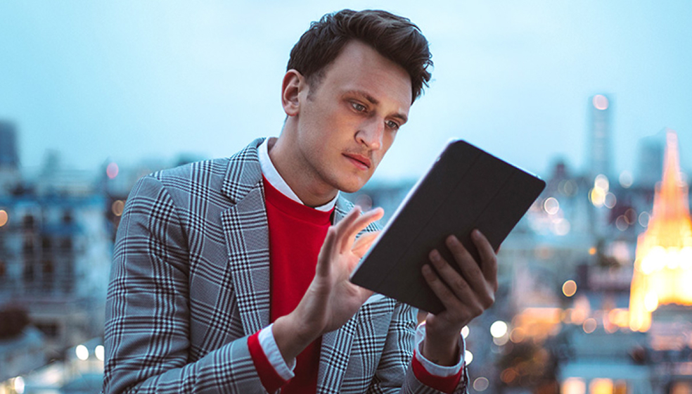 Mann in kariertem Sakko sitzt vor einer City Skyline und tippt auf seinem Tablet