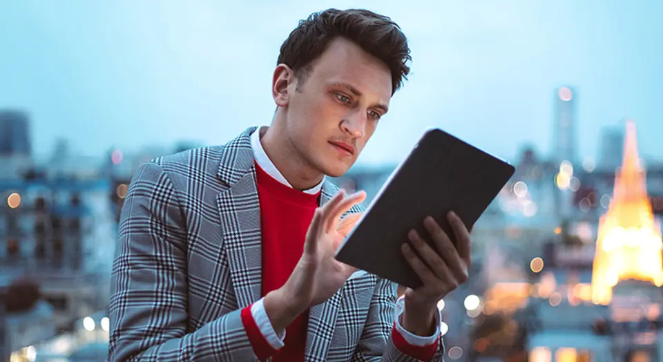 Mann in kariertem Sakko sitzt vor einer City Skyline und tippt auf seinem Tablet