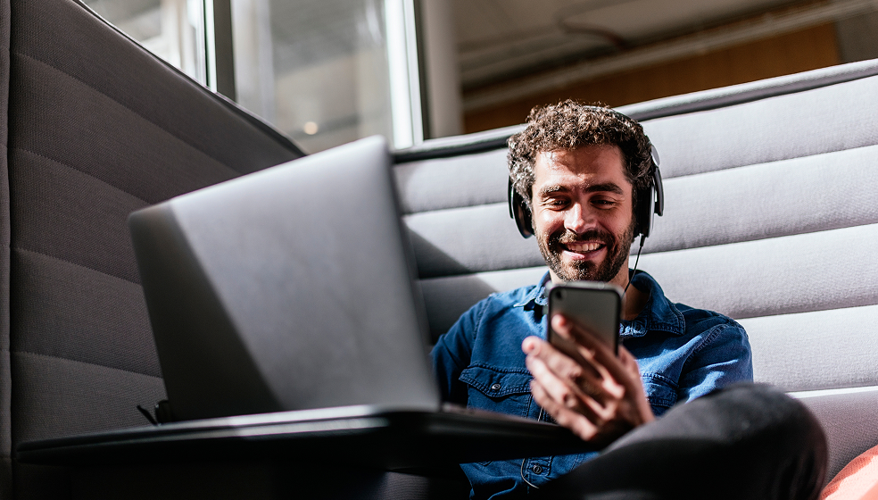 Lockiger Mann mit Headset arbeitet am Laptop und schaut in sein Smartphone