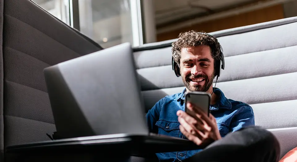 Lockiger Mann mit Headset arbeitet am Laptop und schaut in sein Smartphone