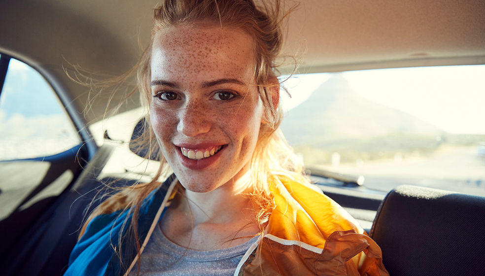 Frau mit Sommersprossen sitzt hinten in einem Auto und lächelt in die Kamera