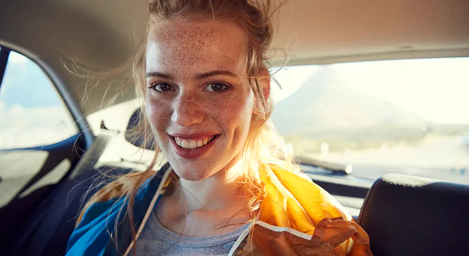 Frau mit Sommersprossen sitzt hinten in einem Auto und lächelt in die Kamera