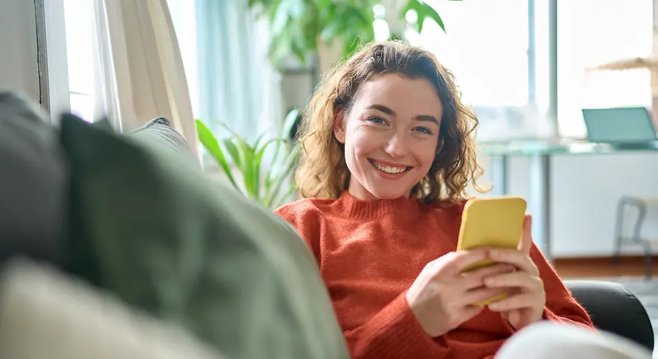 Frau in orangenem Pulli hält gelbes Smartphone in der Hand und lächelt in die Kamera