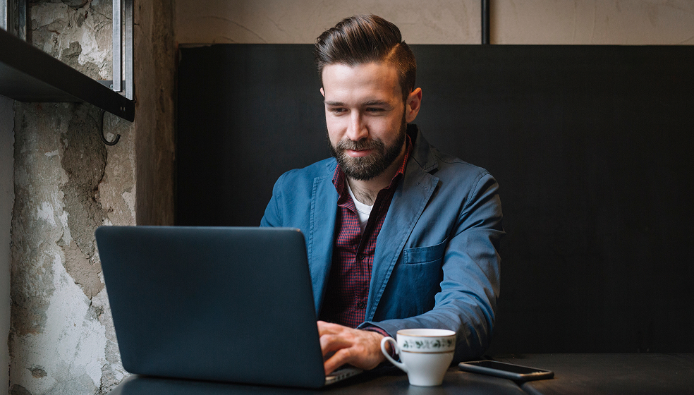 Mann in blauem Sakko sitzt in einem Café und arbeitet an seinem Laptop