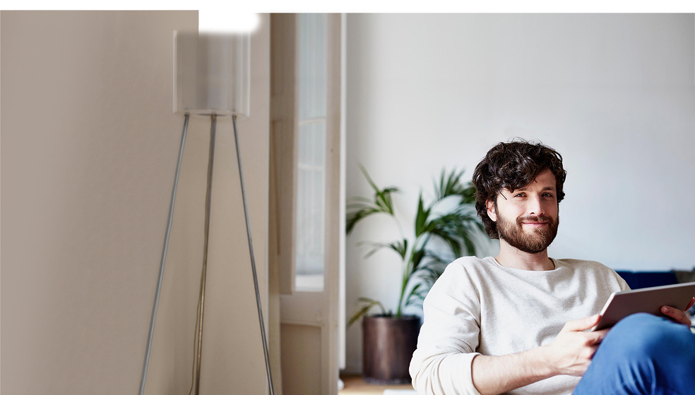 Dunkelhaariger Mann in weißem Pullover sitzt auf einem Sofa und lächelt in die Kamera