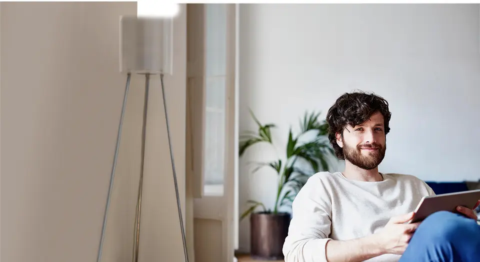 Dunkelhaariger Mann in weißem Pullover sitzt auf einem Sofa und lächelt in die Kamera