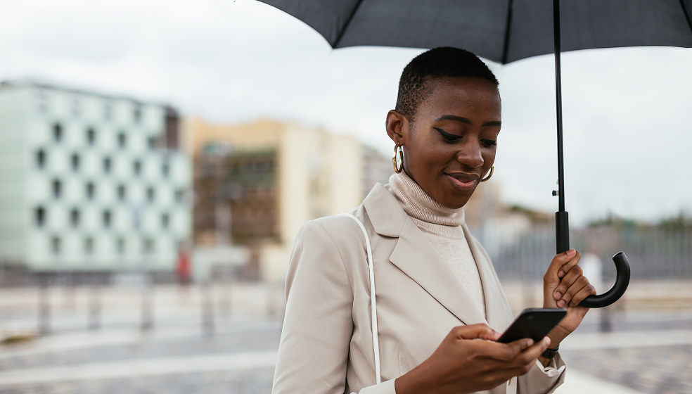 Frau in Mantel mit Regenschirm steht draußen und schaut in ihr Smartphone