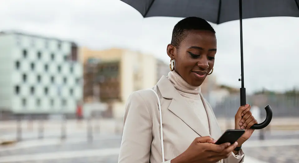 Frau in Mantel mit Regenschirm steht draußen und schaut in ihr Smartphone