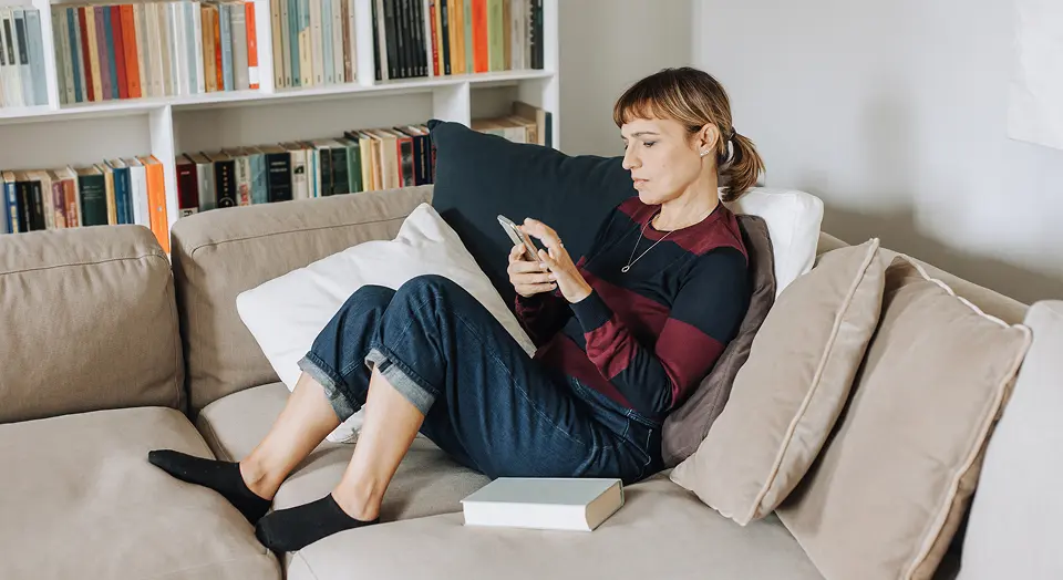 Blonde Frau sitzt in ihrem Wohnzimmer auf dem Sofa und schaut in ihr Smartphone