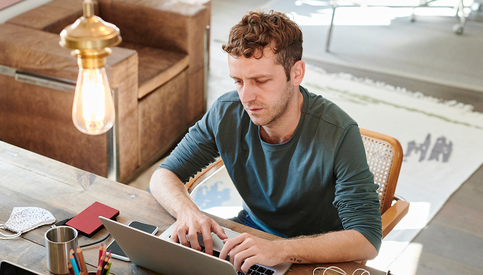 Lockiger Mann sitzt am Schreibtisch und arbeitet an seinem Laptop