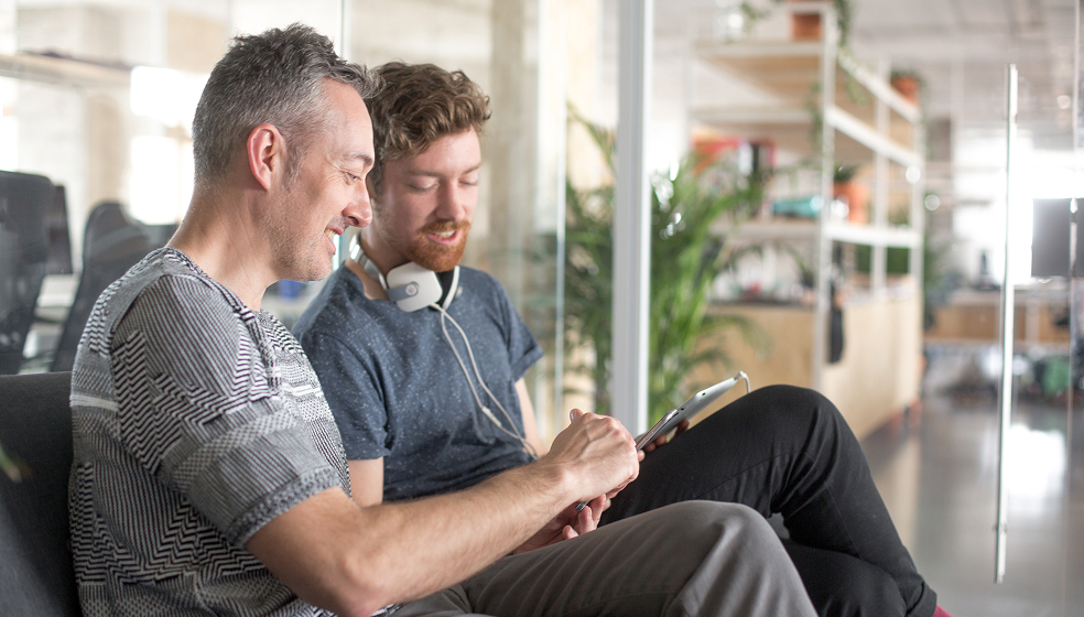 Zwei Männer sitzen im Büro nebeneinander und schauen gemeinsam in ein Tablet
