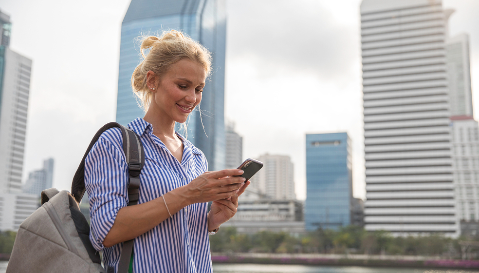 Frau in gestreiftem Hemd steht draußen vor vielen Hochhäusern und schaut in ihr Smartphone