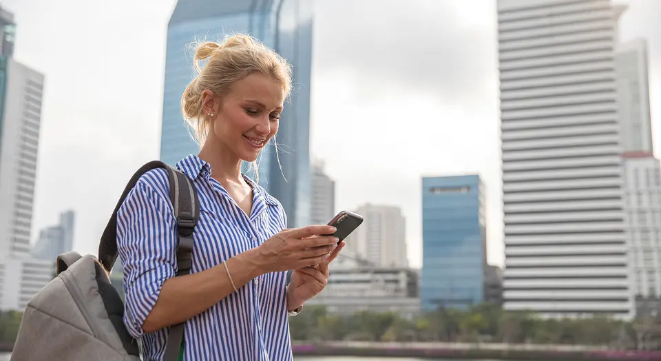 Frau in gestreiftem Hemd steht draußen vor vielen Hochhäusern und schaut in ihr Smartphone