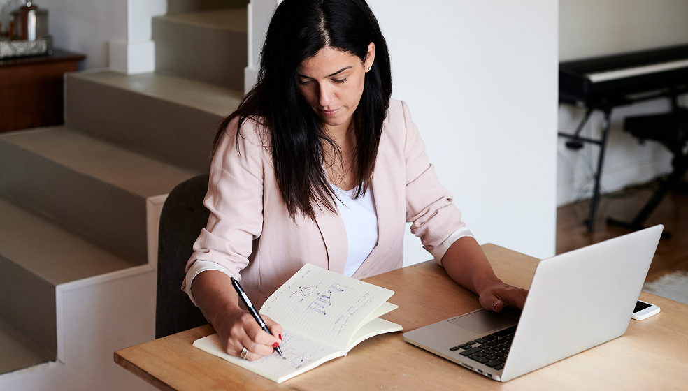 Dunkelhaarige Frau in rosanem Blazer macht ihr Steuererklärung mit Laptop und Notizbuch