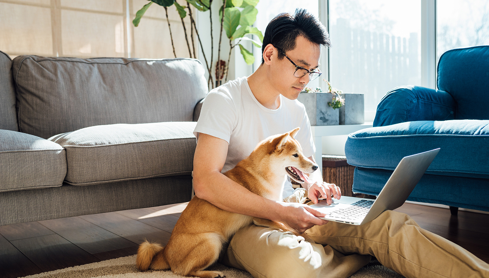 Dunkelhaariger Mann sitzt mit Hund und Laptop auf dem Schoß auf dem Boden vor seinem Sofa