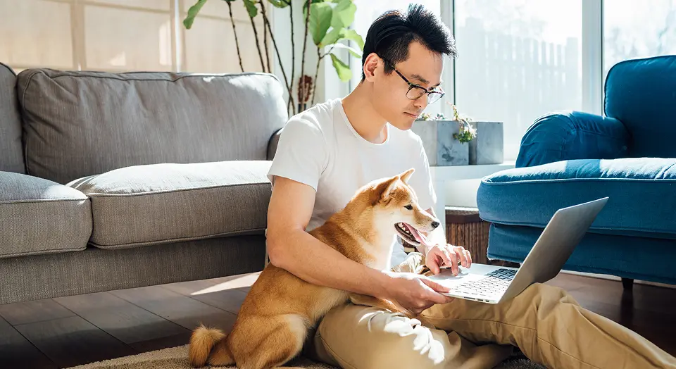 Dunkelhaariger Mann sitzt mit Hund und Laptop auf dem Schoß auf dem Boden vor seinem Sofa