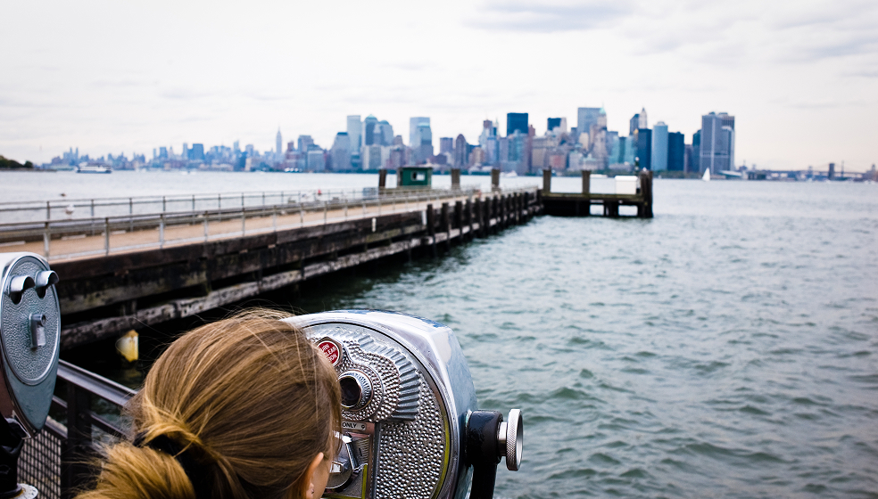 Blonde Frau schaut an einem ufer durch ein Fernglas auf eine City Skyline am anderen Ufer
