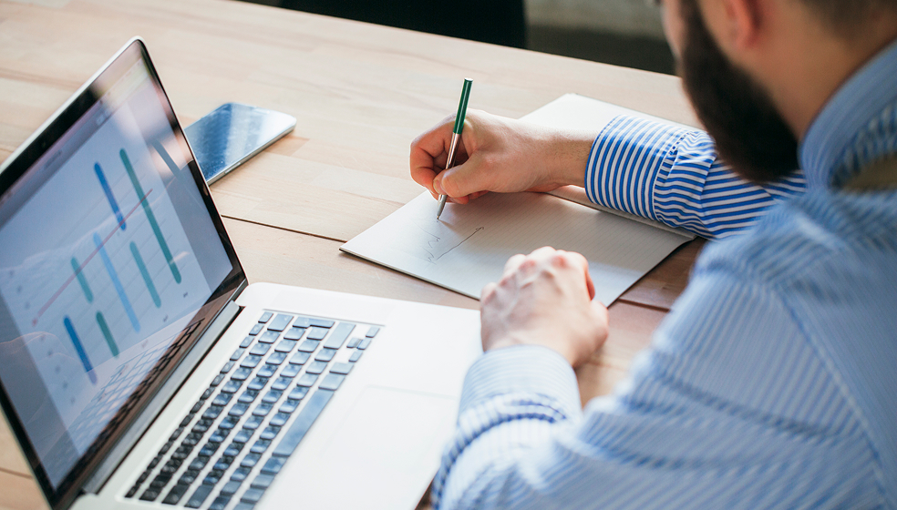 Nahaufnahme Mann in Hemd, der sich neben seinem Laptop auf einem Blatt Papier Notizen macht